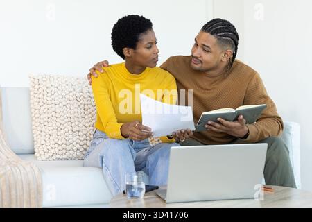 Reviewing documents Diverse couple sitting on sofa in bright living room, with laptop and notebook Stock Photo
