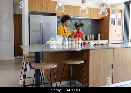 Diverse mother and daughter mixing cupcake batter in home kitchen, with marble island Stock Photo