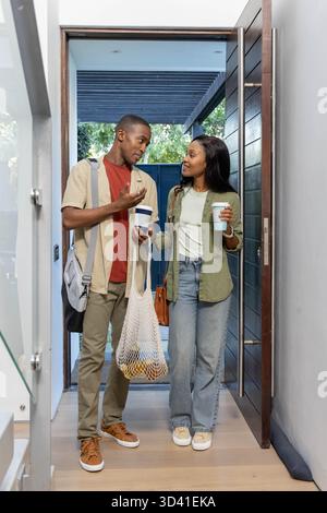 Holding coffee cups and mesh grocery bag, African American couple standing in home entryway Stock Photo