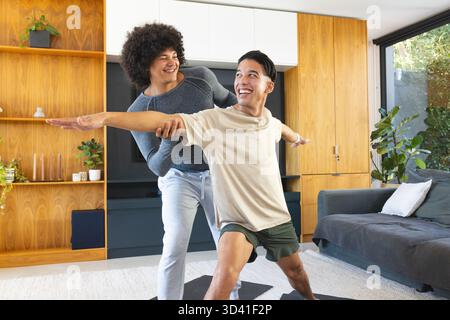 Diverse male friends practicing warrior poses in modern living room, with yoga mats Stock Photo