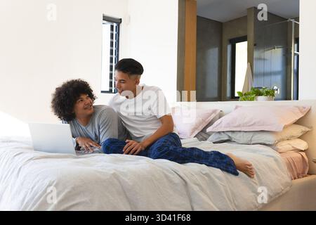 Lounging Diverse male friends using laptop on king-size bed in bright bedroom, with potted plant Stock Photo