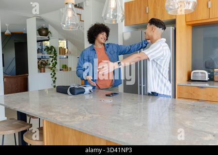 Smiling Diverse male friends reaching out across kitchen island, with marble island, pendant lights Stock Photo