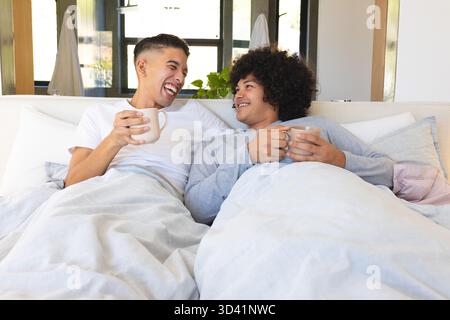 Laughing diverse male partners sitting on bed in bedroom, holding ceramic mugs near potted plant Stock Photo