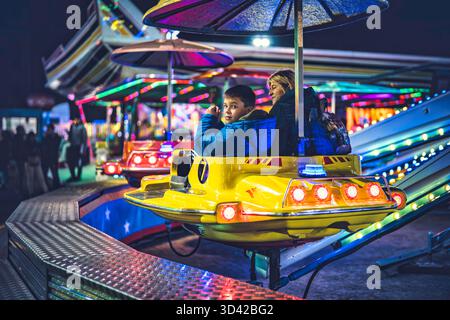 Milan, italy 27 october 2025: family enjoying a colorful carnival ride at night, parents and child smiling under glowing fair lights, festive joyful m Stock Photo