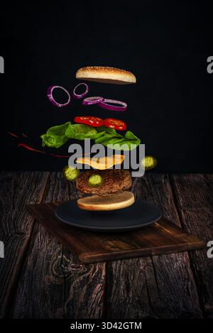 Creative image of floating burger layers with cheese, lettuce, tomato, onions and pickles on rustic wood background. Stock Photo