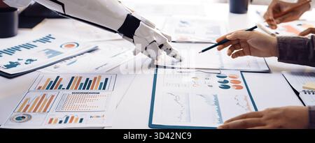 A robotic arm interacts with business analytics documents on a table, showcasing collaboration between technology and human effort in a modern Stock Photo