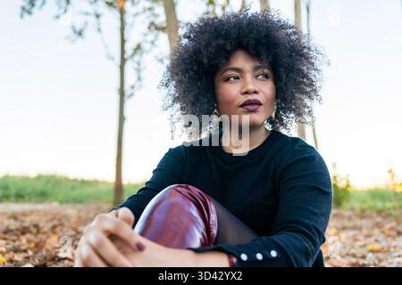 Young black woman with curly hair sitting outdoors looking away with a thoughtful expression Stock Photo