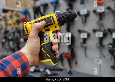 A man in a hardware store chooses a new drill or screwdriver. A hand holds a yellow power drill in front of hardware store display with various tools. Stock Photo