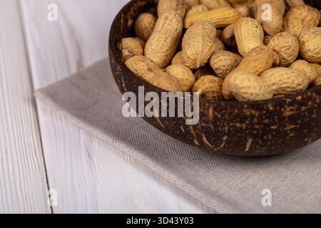 Close-up view of whole peanuts in their shells served in a natural, dark coconut bowl, placed on a beige linen napkin on a white wooden table Stock Photo