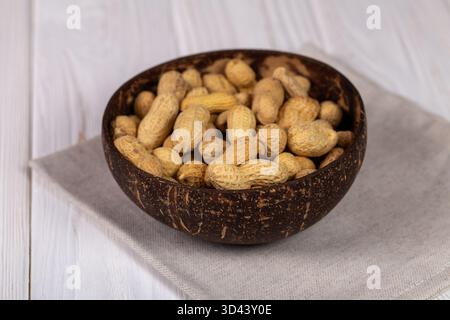 Close-up view of whole peanuts in their shells served in a natural, dark coconut bowl, placed on a beige linen napkin on a white wooden table Stock Photo
