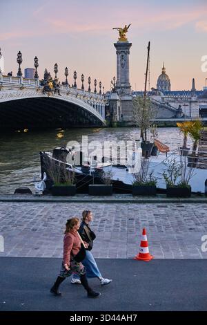 Paris, France - March 2025: Two women walk leisurely by the Seine River as the sun sets, showcasing beautiful Parisian architecture and the glimmering Stock Photo