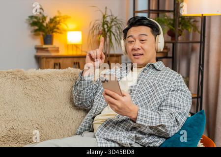 Happy overjoyed middle-aged man in wireless headphones relaxing lying on couch using phone at home choosing, listening favorite energetic disco dancing music. People weekend leisure activities. Stock Photo