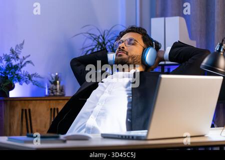Happy smiling Indian businessman listening music on headphones taking break relaxing leaning on chair after working at office workplace. Male Hindu freelancer in formal suit puts hands behind head Stock Photo