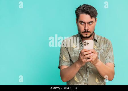 Middle-aged man anxiously types fast on smartphone, breathing deeply while focused on composing urgent message. Young adult guy on blue background shows tension, nervousness, digital communication. Stock Photo