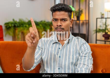 Indian man at home waves finger no no to camera and shows refusal and disagreement with stern mood. Arabian guy on sofa shakes head denies request points finger and shows unhappy sad reject reaction Stock Photo