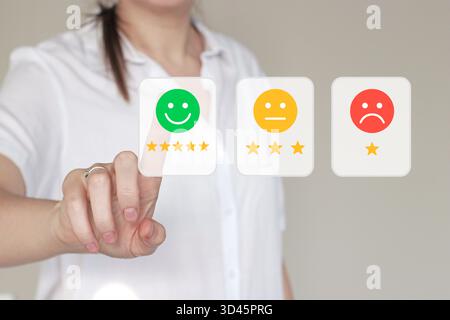 Female hand selecting a smiley face rating on transparent digital screen. Customer review, satisfaction level, and quality control in modern business Stock Photo