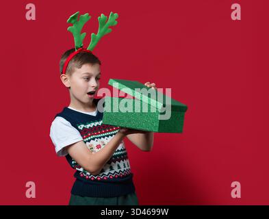 A surprised boy in holiday sweater and green reindeer antlers opening a green glitter Christmas gift box, red background Stock Photo
