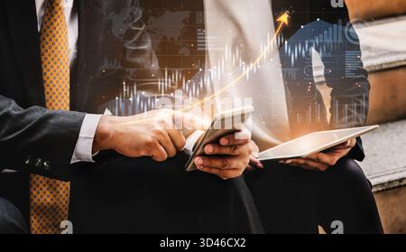 Two business professionals engage in a discussion on financial growth, utilizing technology like a smartphone and tablet, with an overlay of market Stock Photo