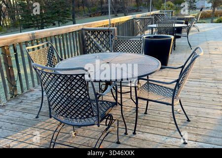 Empty metal patio tables and chairs on a wooden deck in morning sunlight, outdoor seating area surrounded by trees and nature. patio furniture on wood Stock Photo