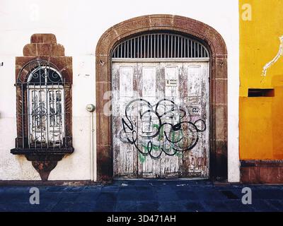 Weathered wooden door with graffiti and arched stone frame next to an old iron-barred window on a colorful urban building facade Stock Photo