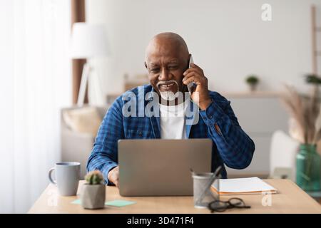Mature African Man Using Laptop and Talking on Cellphone in Modern Office Setting Stock Photo