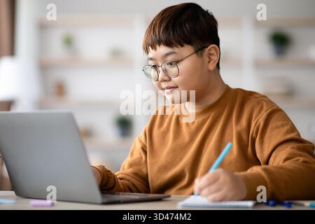 Plump Teenager With Glasses Enjoys Doing Homework on Laptop While Taking Notes at Home Stock Photo