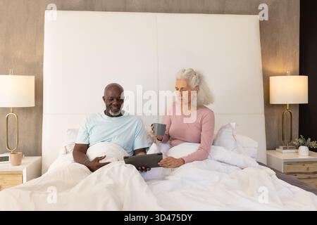 Reading senior couple using tablet computer in bedroom under white bedding, with coffee mug Stock Photo
