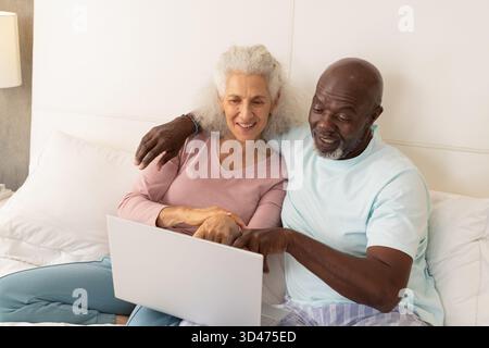 Smiling diverse senior couple leaning against white headboard in cozy bedroom, using silver laptop Stock Photo