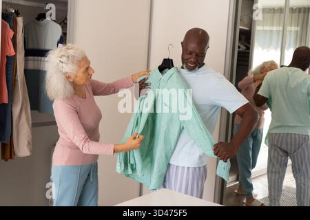 Holding Diverse couple trying on light green shirt in bedroom area, with wardrobe and mirror Stock Photo