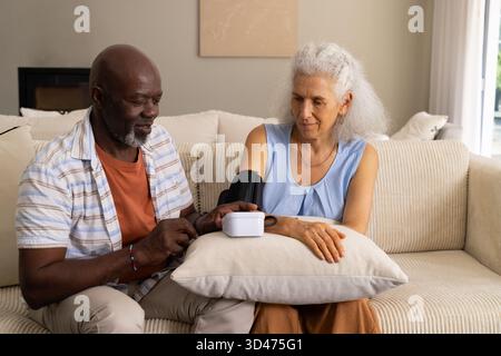 Measuring blood pressure for Diverse senior couple on sofa at home, using cuff and monitor Stock Photo