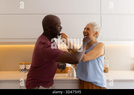 Diverse senior couple sharing feeding moment in home kitchen, with bowl and stone countertop Stock Photo
