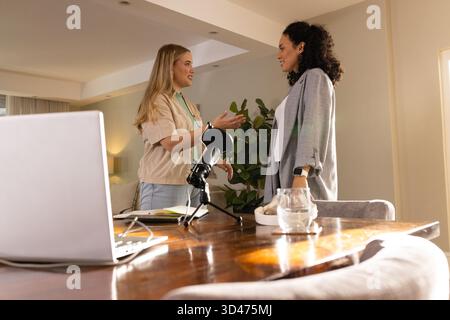 Talking Diverse female colleagues standing across table in living room, with podcast microphone Stock Photo