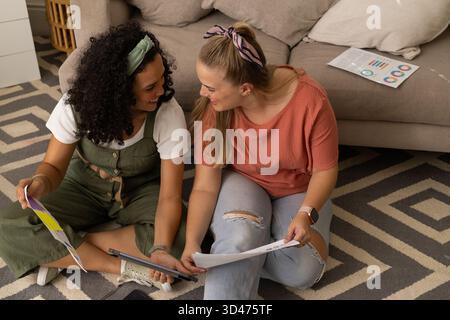 Collaborating diverse female friends reviewing charts and graphs on rug in living room, with tablet Stock Photo