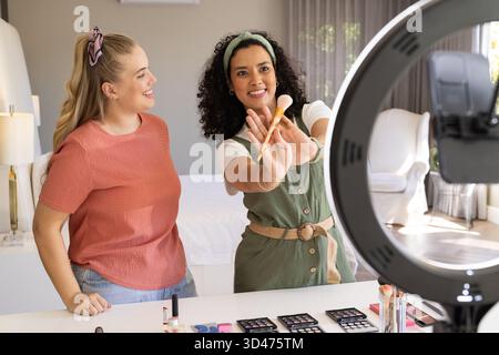 Presenting diverse female friends holding makeup brushes on white desk in bedroom, with ring light Stock Photo