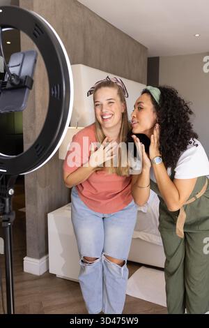 Smiling Diverse female friends leaning toward ring light in modern bedroom, with mounted smartphone Stock Photo
