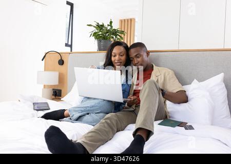 Leaning close African American couple studying on white bed in bedroom, with laptop and passport Stock Photo