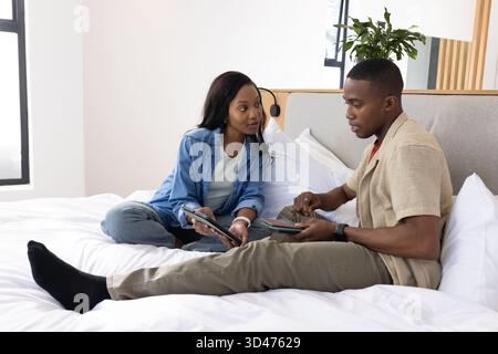 Collaborating African American couple sitting on bed in modern bedroom, using tablets and styluses Stock Photo