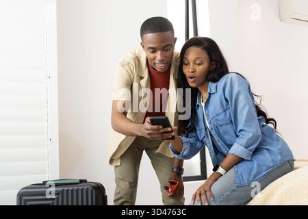 Leaning forward African American couple checking smartphone in bedroom, with suitcase and tablet Stock Photo