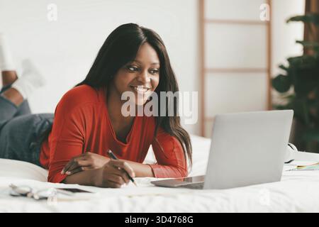 Smiling Woman Studies Online on Her Laptop While Lying on a Bed at Home Stock Photo