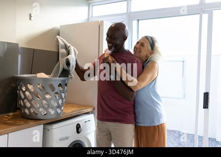 Laughing Diverse couple hugging and folding laundry in laundry room, with washing machine Stock Photo