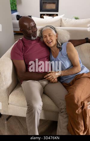 Sitting diverse senior couple holding hands on cream corduroy sofa in living room, with fireplace Stock Photo