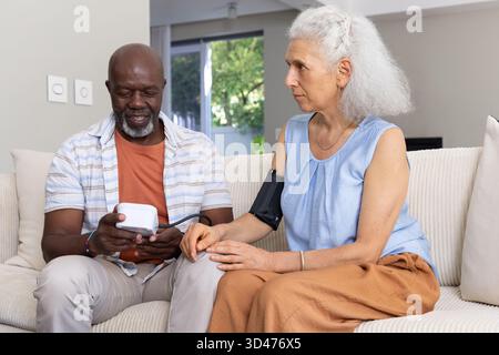 Measuring Diverse senior couple sitting on sofa in living room, using digital monitor and cuff Stock Photo
