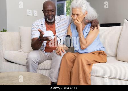 Diverse senior couple holding digital blood pressure monitor on couch in room, with surprised face Stock Photo