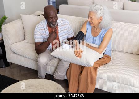 Monitoring diverse senior couple using blood pressure monitor in living room, with pillow Stock Photo