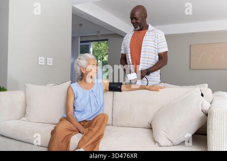 Measuring diverse senior couple using blood pressure cuff on sofa in living room, with monitor Stock Photo
