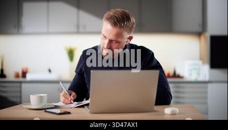 Young Caucasian Man Attending Online Course In His Domestic Kitchen. Writing Essay, Listening To Music Stock Photo