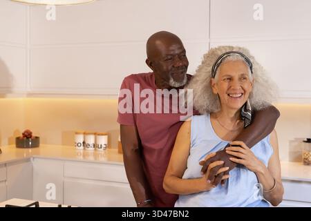 Hugging Diverse senior couple standing in modern home kitchen, with potted succulent and cereal jar Stock Photo