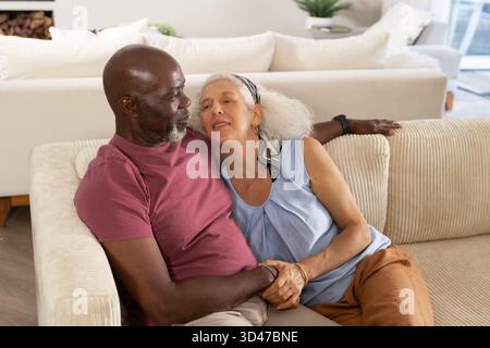 Holding hands diverse senior couple leaning together on sofa in living room, with firewood stack Stock Photo