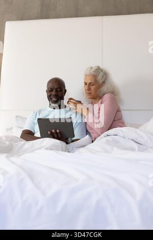 Using tablet Diverse senior couple relaxing in bed at home, with ceramic mug and white bedding Stock Photo