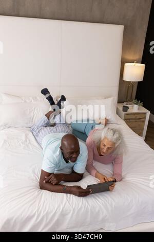 Lying Diverse senior couple using tablet on bed with white linens in bedroom, with bedside lamp Stock Photo